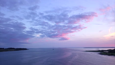 aerial shot flying out over a calm atlantic ocean harbor with a single sailboat during a colorful pink and purple sunset off the maine coast