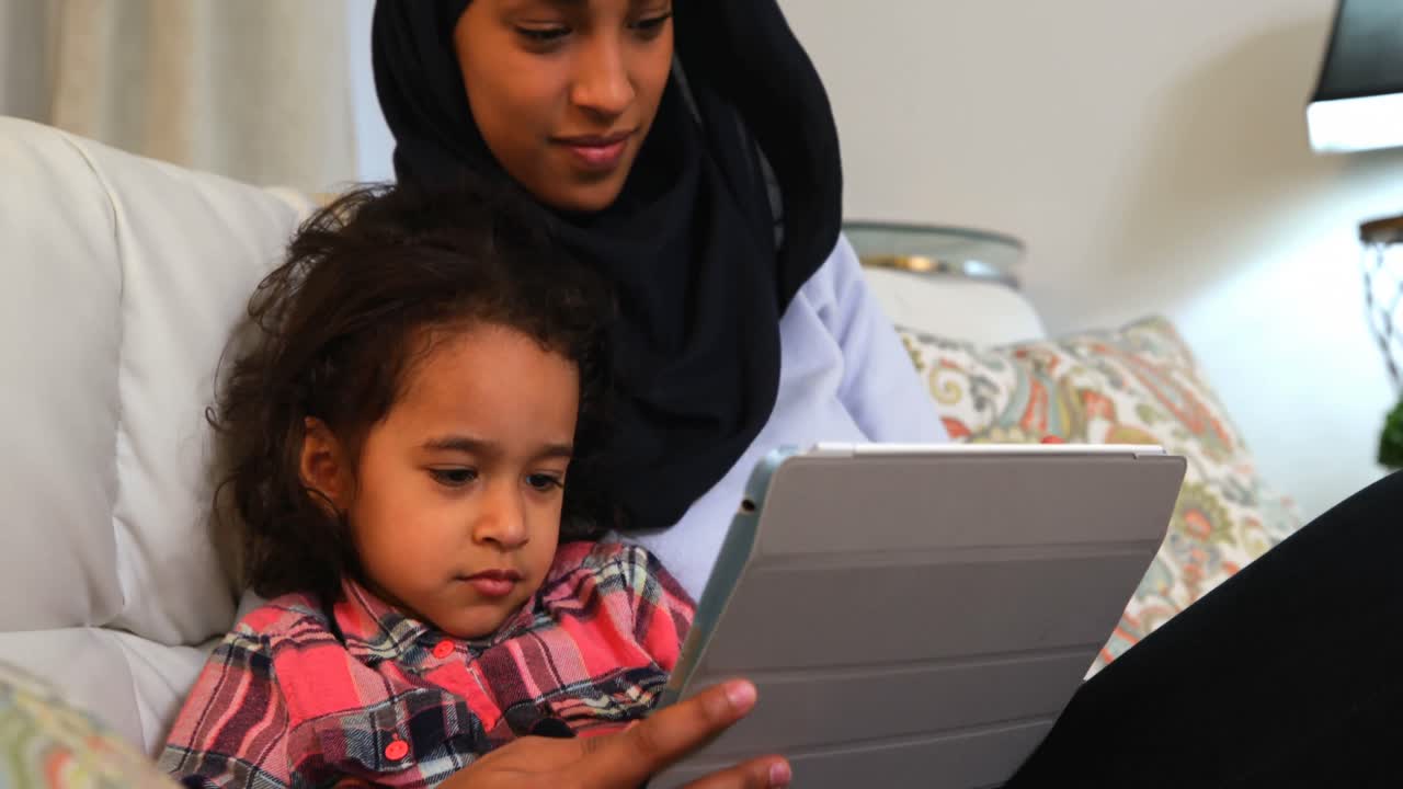Young mother using a digital tablet with her daughter on the sofa at home 4k