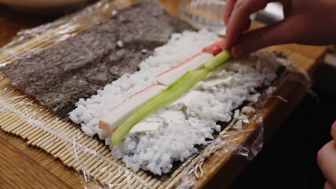 persona colocando pepino junto al cangrejo sobre arroz en rollo de sushi, preparación casera