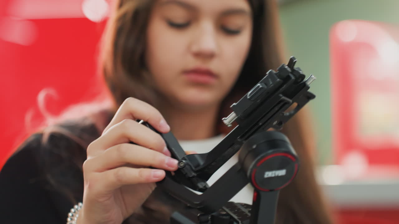white student calibrating gimbal closeup, hands tuning mount and screws, headphones around neck, red bokeh cafe background, focused teenage technician testing rig for vlog, precision adjustments