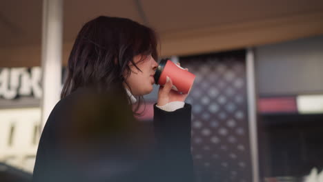 primer plano de una mujer con abrigo negro bebiendo café de una taza roja al aire libre, colocándolo suavemente en satisfacción mientras se centra en la computadora portátil, con un fondo urbano borroso