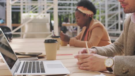 Hombre-De-Negocios-Con-Ropa-Elegante-Sentado-En-La-Terraza-Del-Bar-Mientras-Habla-Por-Teléfono-Con-Auriculares,-Mujer-Comiendo-En-El-Fondo