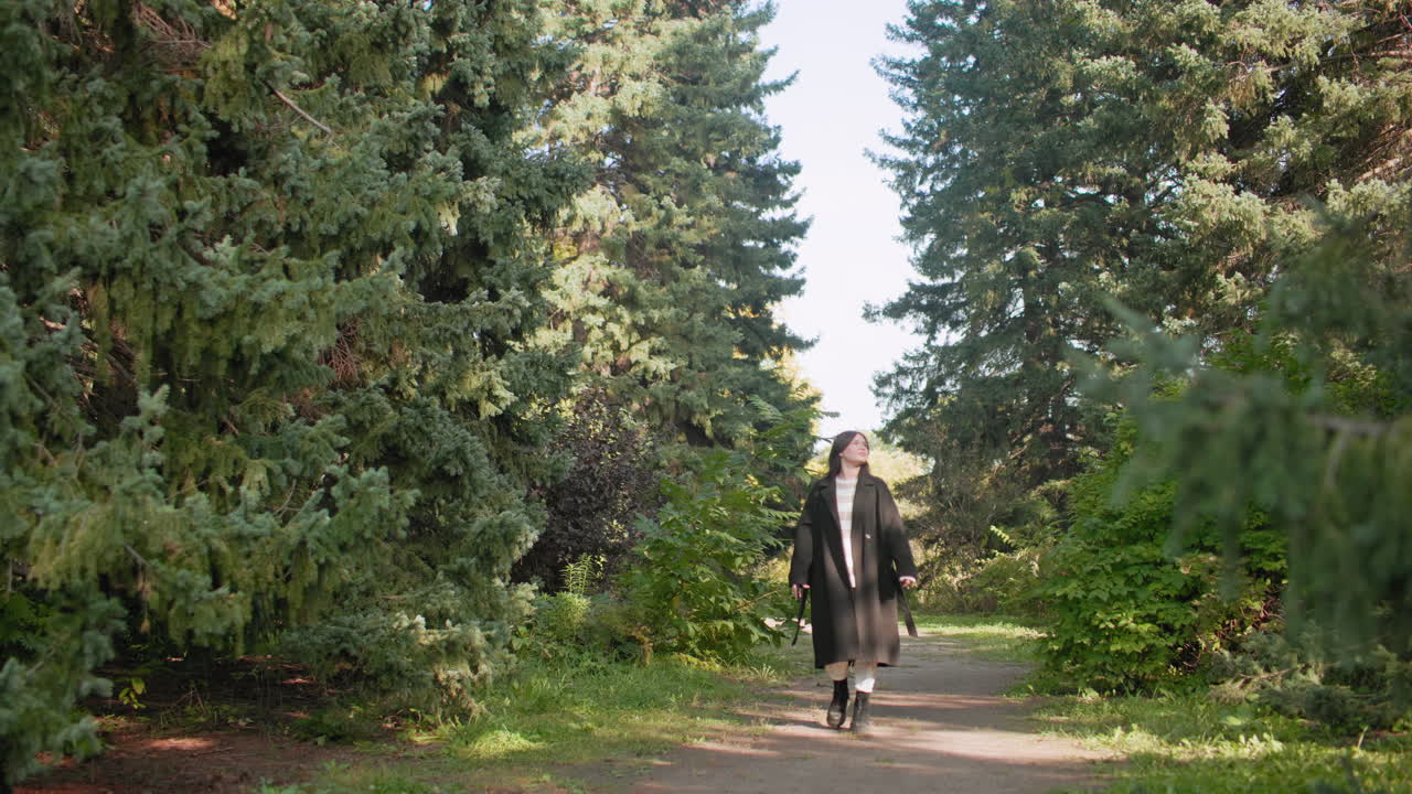 가을 유모차를 끌고 긴 겨울 코트를 입은 사람이 수목원을 거닐며 휴대폰을 들고 사진을 찍고 있어요. 우뚝 솟은 상록수가 고요한 길을 감싸고, 부츠는 오솔길에서 바삭거리는 소리를 내며 평화로운 자연 속을 거닐고 있습니다.