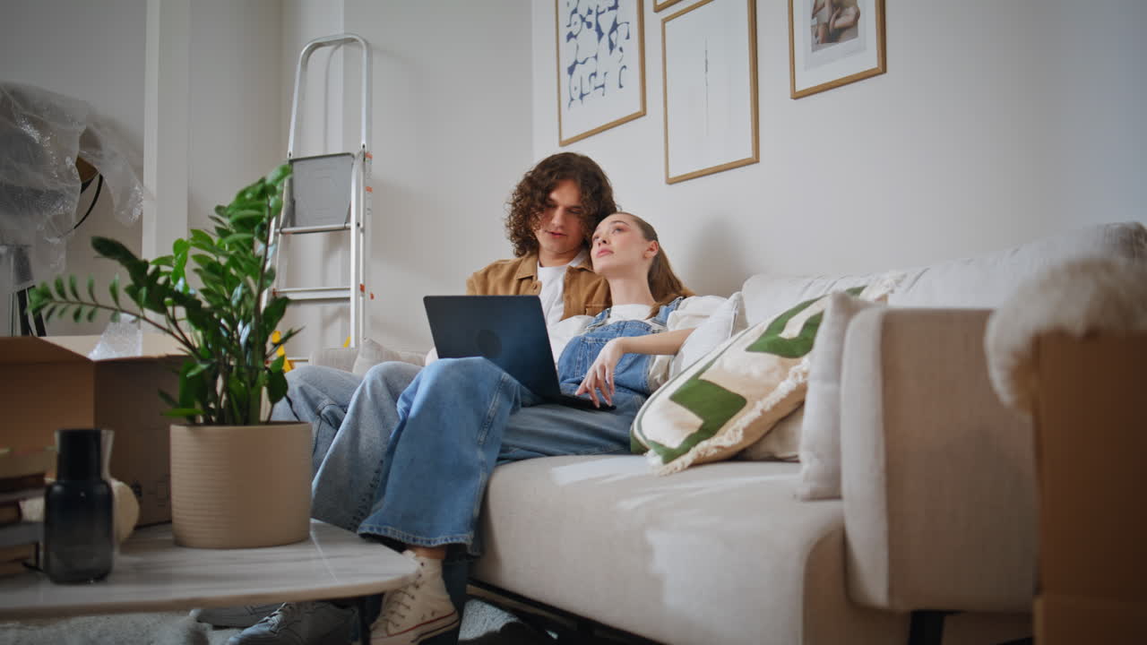Loving spouses sitting sofa new home with laptop. Calm woman resting with man