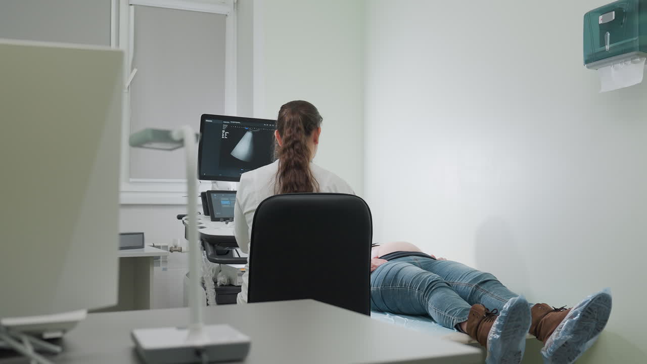 Lab scientist seated in front of diagnostic machine attending to patient lying on bed during examination inside clean clinical room with desktop computer, ultrasound monitor, and lamp stand