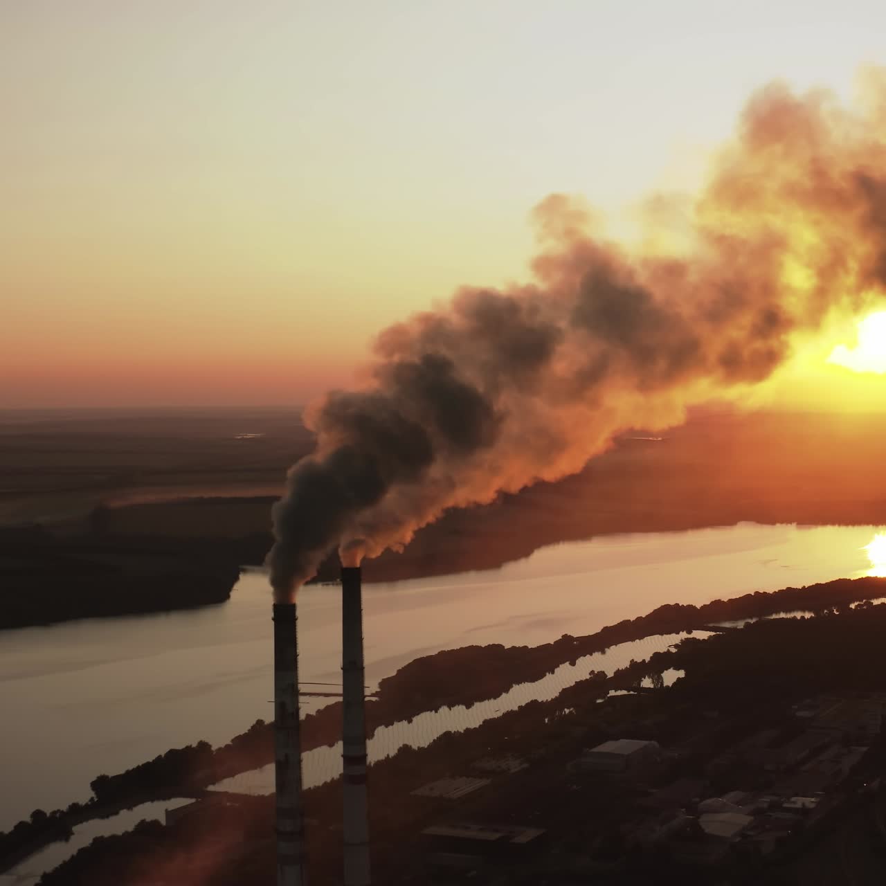 Massive air pollution near the river at darkness. Huge puffs of harmful smoke exhaust into the air against setting sun. Chimneys with dark fumes from manufacture. Aerial view