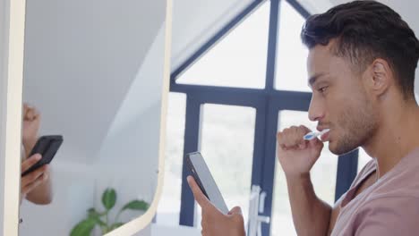 Hombre-Birracial-Cepillándose-Los-Dientes-En-Un-Baño-Soleado-Por-La-Mañana-Y-Usando-Un-Teléfono-Inteligente,-Cámara-Lenta