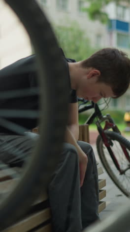 primer plano de la rueda giratoria de la bicicleta en un enfoque agudo, creando un efecto borroso de alguien sentado en un banco con la cabeza inclinada hacia abajo, el fondo presenta edificios, vegetación y árboles