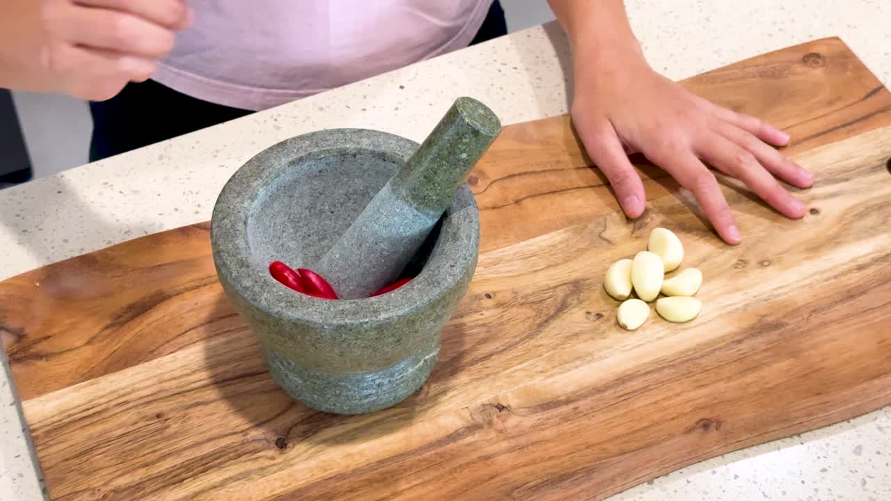 Hands add garlic cloves and red chilies to a stone mortar, then crush them using a pestle on a wooden cutting board under bright kitchen lighting