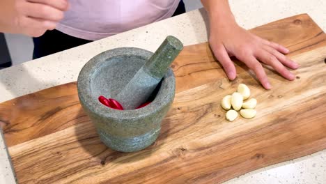 hands add garlic cloves and red chilies to a stone mortar, then crush them using a pestle on a wooden cutting board under bright kitchen lighting
