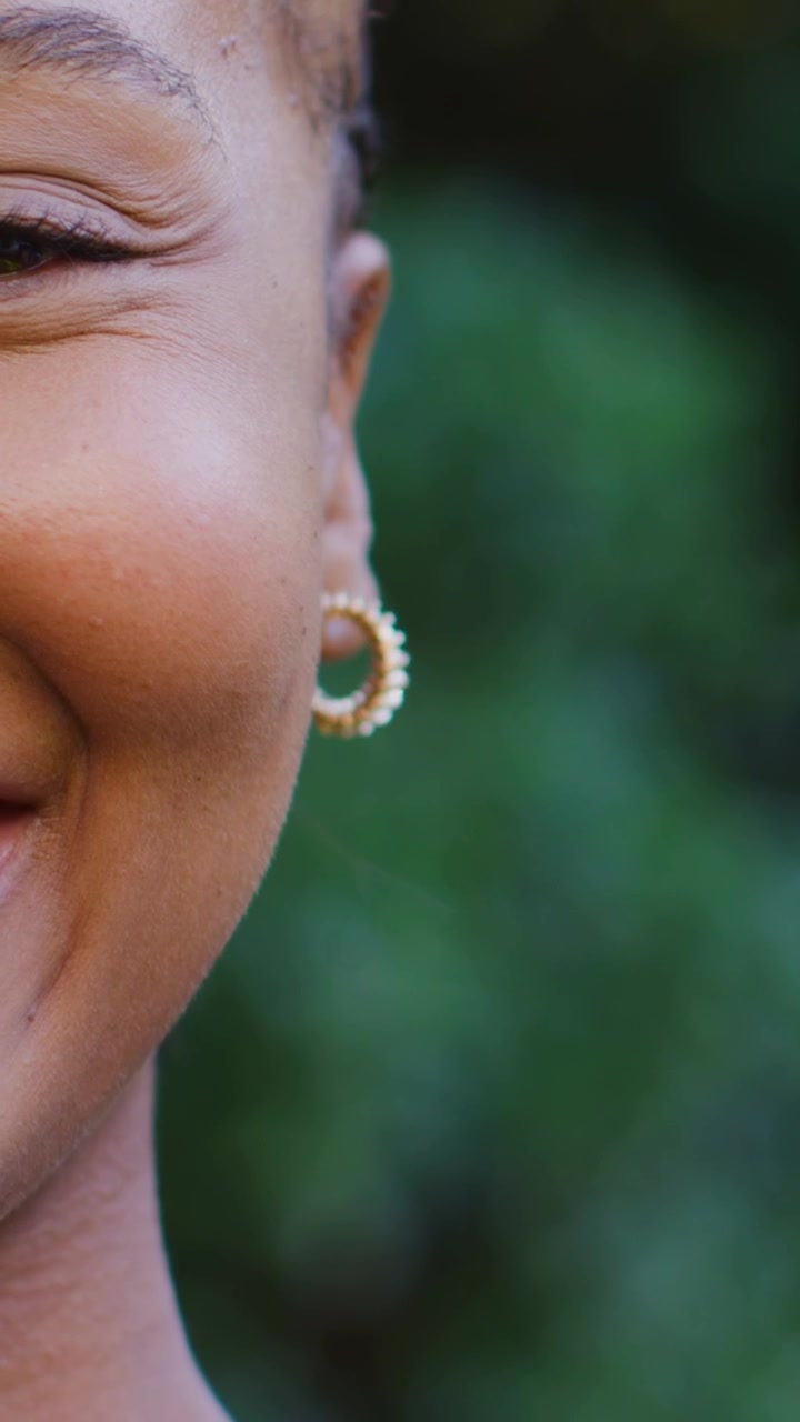 Video verticale: La telecamera inquadra un adolescente che mantiene il contatto visivo, sorridendo con bokeh verde, con un orecchino a cerchio d'oro.