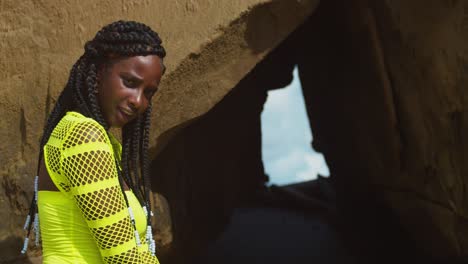 dark skin african girl kneeling in the sand in front of a cave with clouds in the background