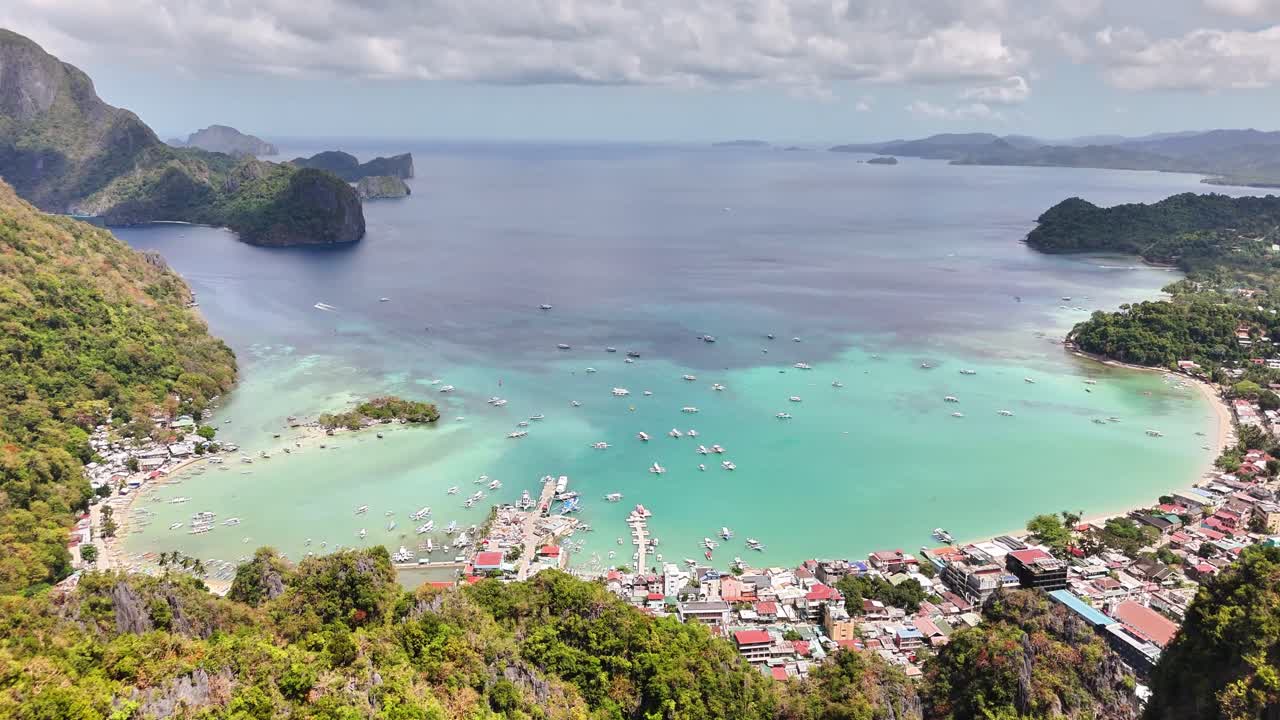 Aerial view shows El Nido’s crescent-shaped bay with boats floating on clear aquamarine waters, framed by limestone cliffs and lush tropical vegetation on northern Palawan’s coast