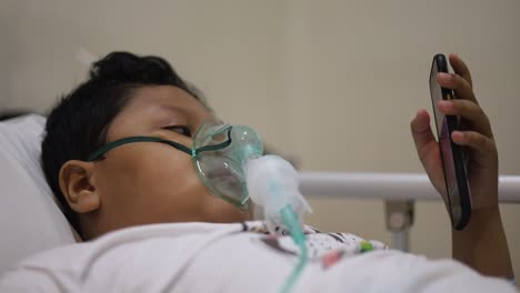 asian 9 years old boy with oxygen mask on her face lie down on bed in the hospital while playing cell phone