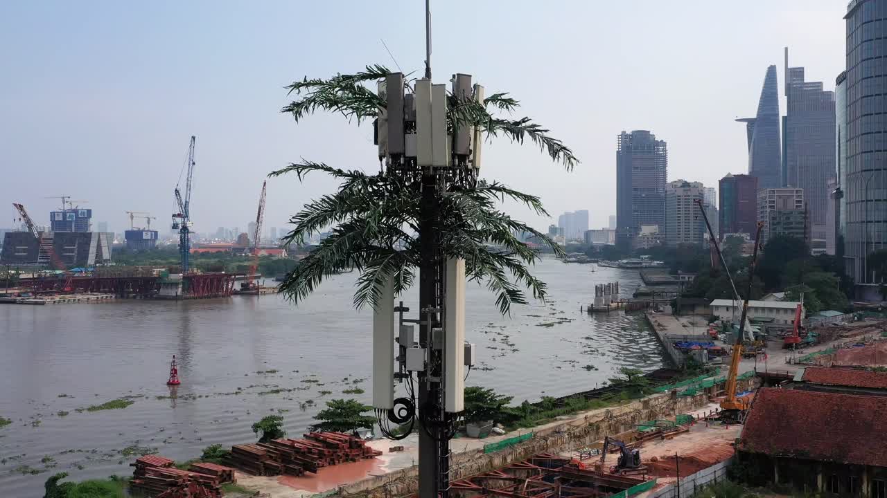 toma aérea descendente de una torre de telefonía móvil ultramoderna disfrazada de palmera contra el fondo de un sitio de construcción frente al río con edificios de la ciudad, grúas y tráfico de barcos en el río