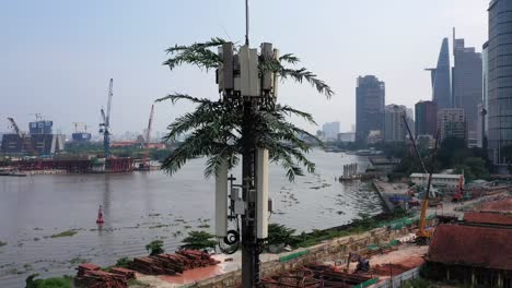 descending-aerial-shot-of-ultra-modern-mobile-phone-tower-disguised-as-a-palm-tree-against-a-background-of-a-riverfront-construction-site-with-city-buildings,-cranes-and-boat-traffic-on-the-river