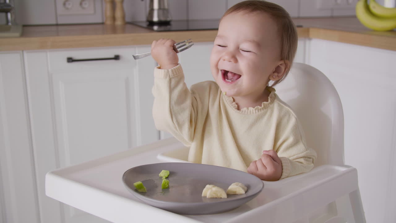 Cute baby girl eating avocado slice using fork while sitting in high chair in the kitchen