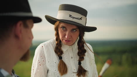 a close-up captures the intense expression of a woman in a white dress, with braided hair and a wide-brimmed hat, as she gives a cool, discerning look toward a painter . she holds a paintbrush