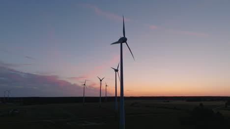 aerial establishing view wind turbines generating renewable energy in a wind farm, evening after the sunset golden hour, countryside landscape, high contrast silhouettes, orbiting drone shot