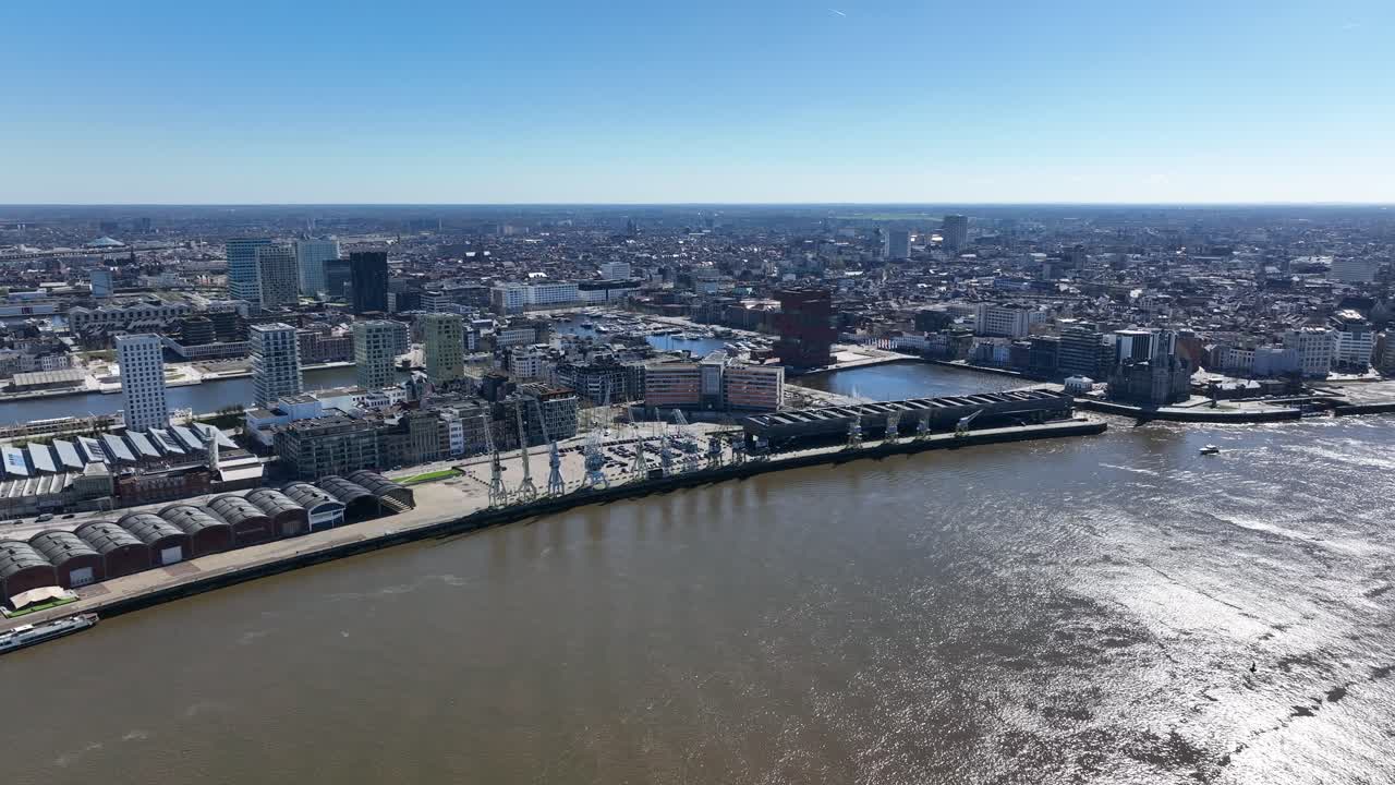Pan left aerial shot of Antwerp skyline with Scheldt river, harbor docks, and cranes on a bright sunny day