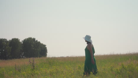 woman in green dress and wide white hat strolls peacefully through tall grassy field beneath clear sky, surrounded by wildflowers and warm sunlight