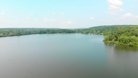 slow-rotating-drone-flight-over-a-lake-in-a-forest-with-people-kayaking-under-a-blue-summer-sky-with-white-clouds-4k