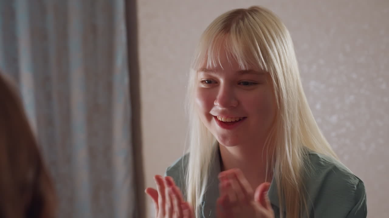 Close up of smiling blonde girl clapping hands playfully with unseen partner in cozy room, showing joy and connection during relaxed indoor bonding moment with curtain backdrop and soft lighting