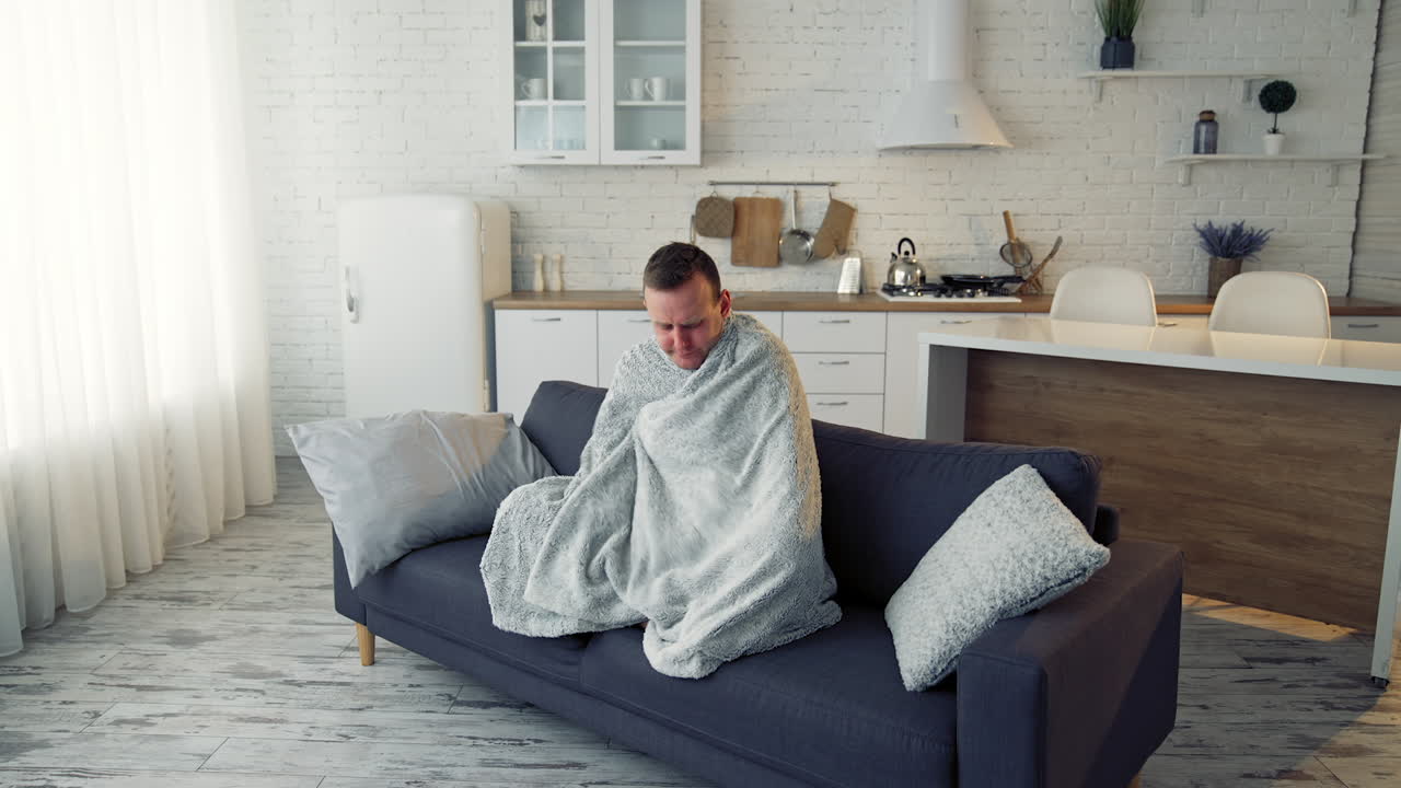 Man having fever. Young man feeling freezy and covering with warm blanket while sitting on a couch at home. Sick man wrapped with blanket during covid outbreak.