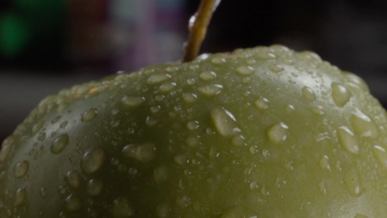 Macro primo piano di una mela verde coperta di goccioline d'acqua, con frutta fresca, pelle liscia, texture naturale e un gambo con umidità sulla superficie.