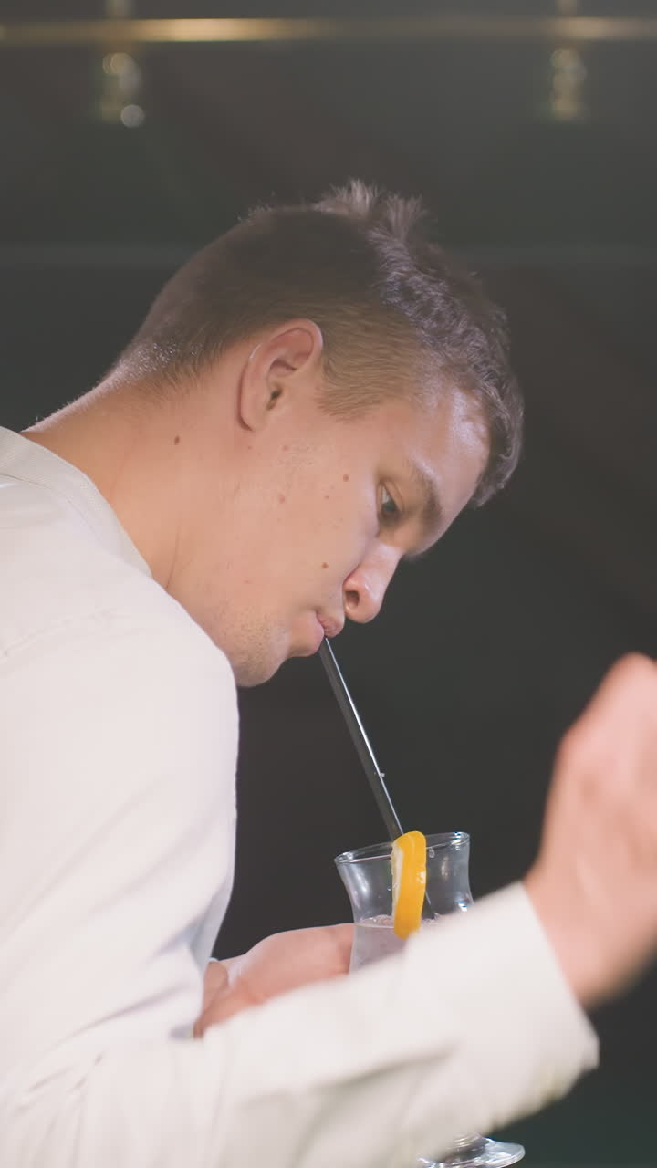 primer plano de un joven con camisa blanca sosteniendo un vaso de bebida de limón con paja negra mientras bebe. la iluminación ambiental destaca la atmósfera relajada, creando un entorno moderno y elegante