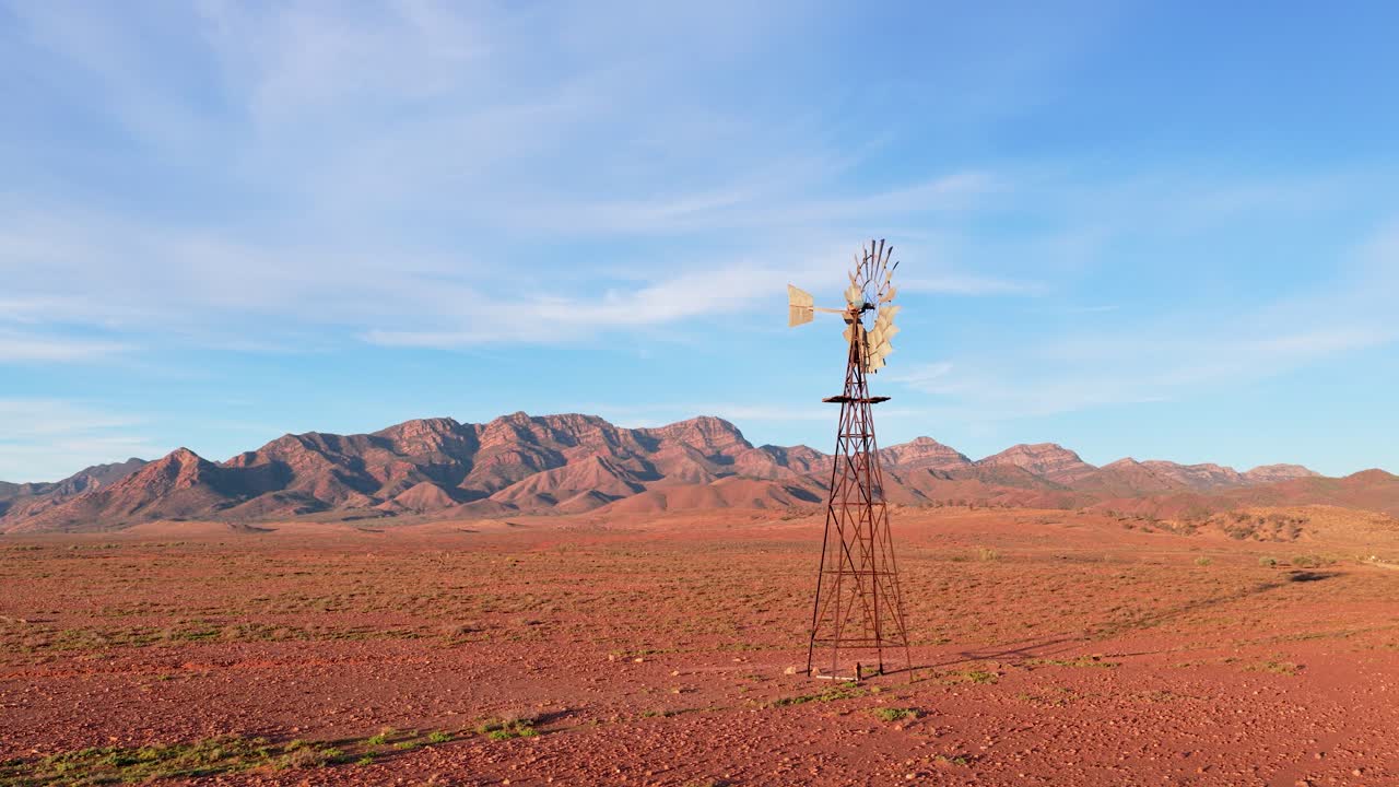 Órbita de dron alrededor de un molino de viento con las escarpadas montañas de Flinders Ranges al fondo en el interior rojo, Australia Meridional