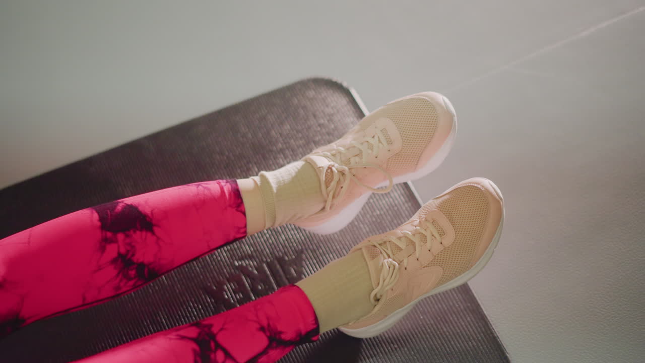 Leggings rosa su tappetino che allungano le gambe, primo piano della parte inferiore del corpo che esegue un sollevamento gambe da seduti su un tappetino scuro da palestra, scarpe da ginnastica beige visibili, illuminazione soffusa dall'alto, atmosfera calma di riscaldamento, prova di allenamento personale.