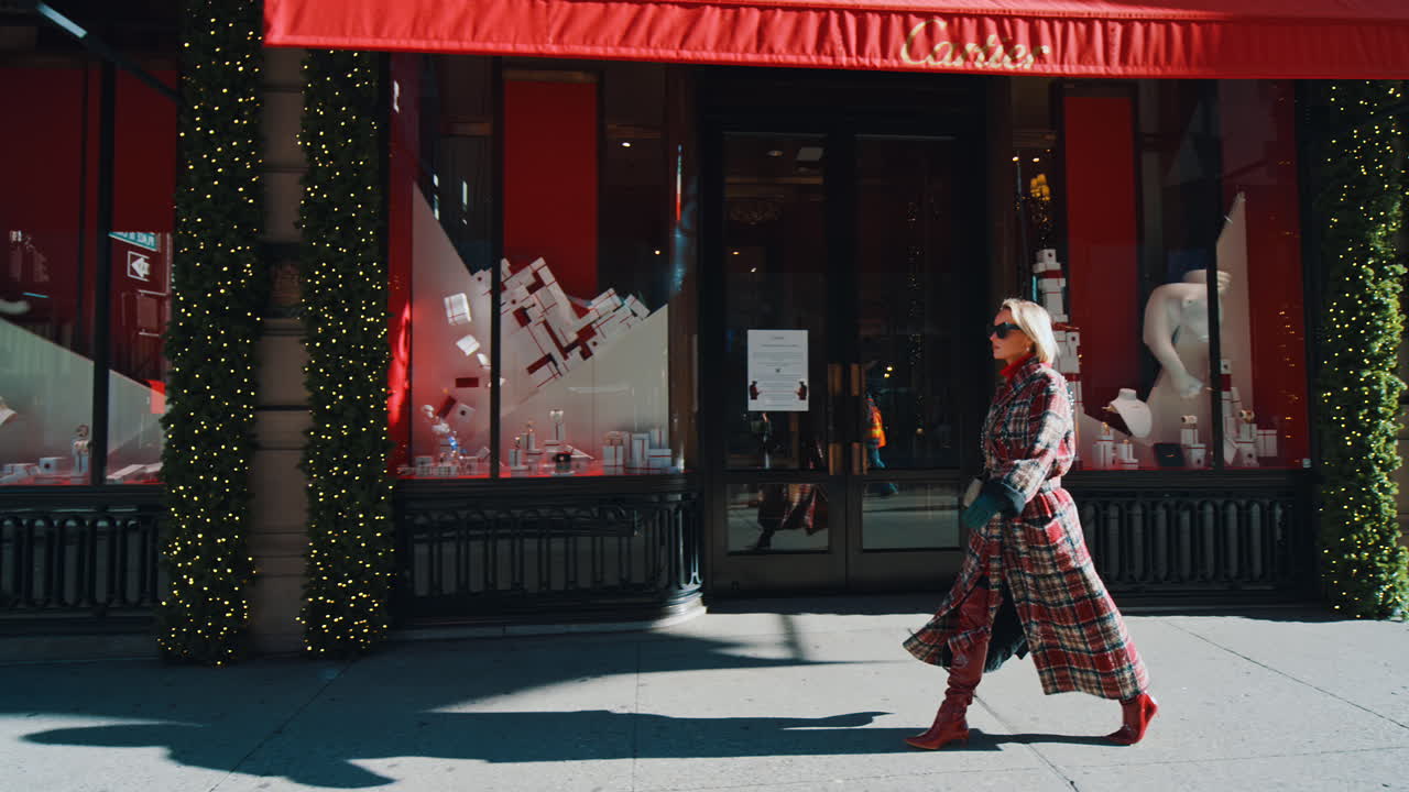 New York, USA, 25 ottobre 2025: Donna in cappotto a quadri che cammina davanti alla vetrina di un negozio di lusso. Veduta laterale di una donna che cammina davanti a una boutique con decorazioni rosse e ghirlande sempreverdi a New York.