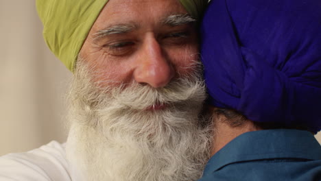 two sikh men embrace each other affectionately, with the older man smiling warmly and the younger man looking down.