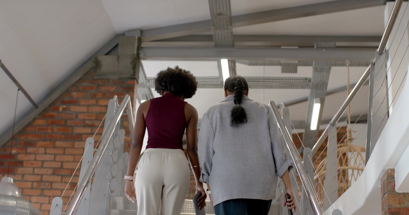 Collaboratrices diverses montant des escaliers métalliques, l'une avec un téléphone bordeaux, se dirigeant vers l'étage.