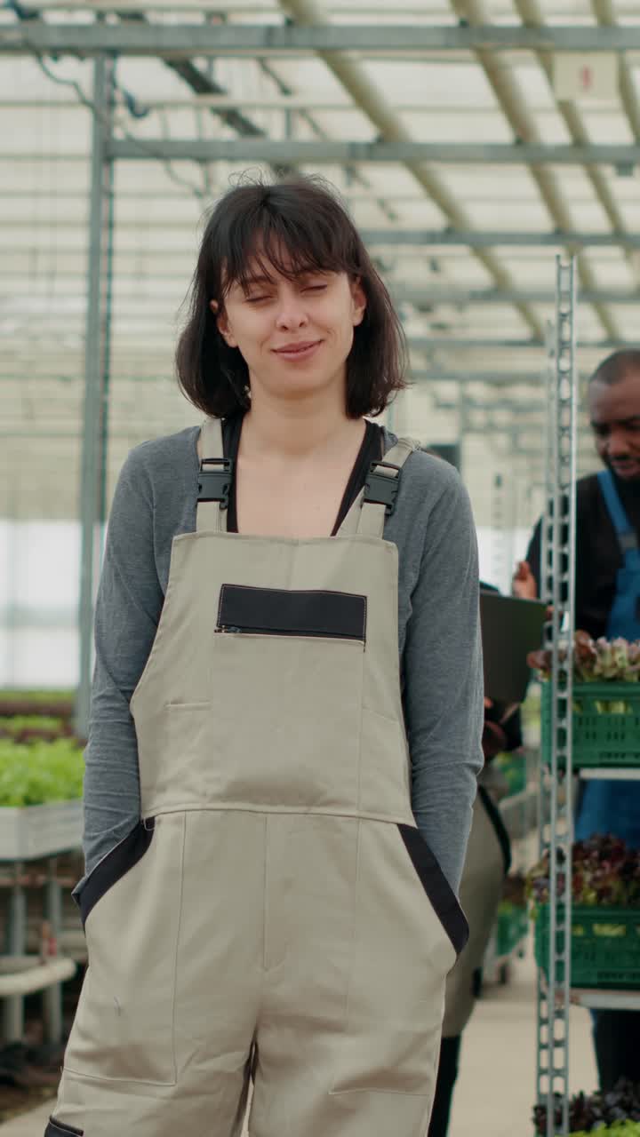 Ritratto di una donna sicura di sé che posa con le mani in tasca in una serra, mentre dei braccianti agricoli usano un laptop