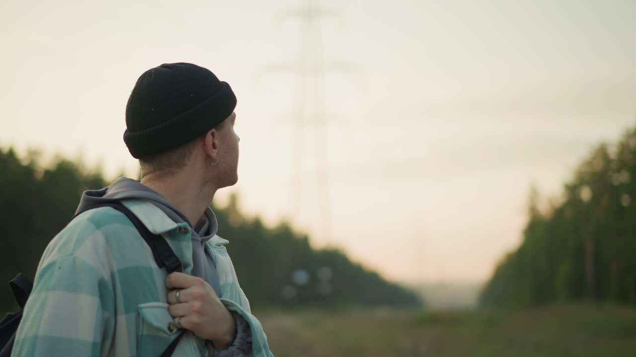 Escursionista bianco che guarda un traliccio in una radura al crepuscolo, viaggiatore pensieroso che stringe la cinghia dello zaino mentre osserva lo skyline e le sfumature del tramonto, un corridoio di campi silenzioso evoca riflessione e percorso