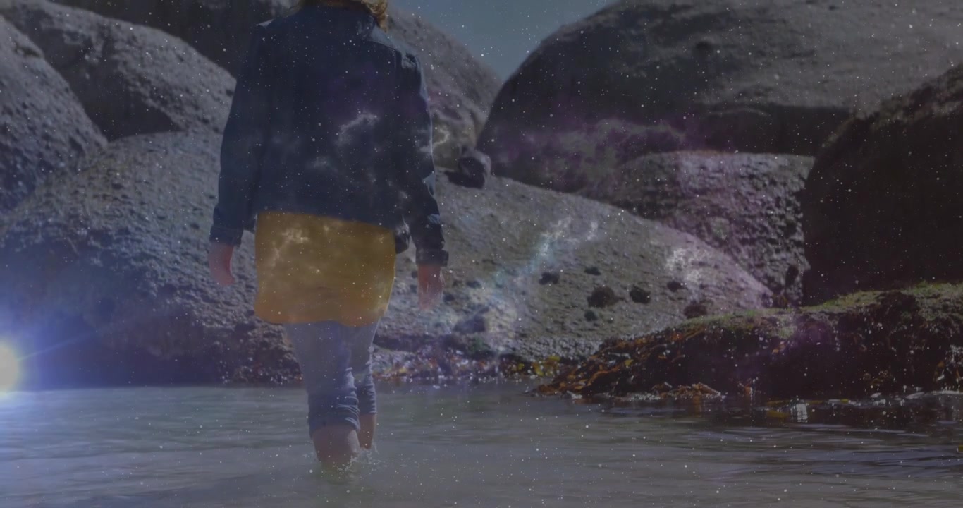 Donna che entra in una pozza di marea, dirigendosi verso le rocce, esplorando mentre un bagliore dell'obiettivo traccia le increspature.