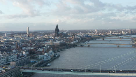 Aerial-shot-of-Rhine-river-calmly-flowing-through-city.-Backwards-reveal-of-modern-busy-multilane-road-bridge.-Cologne,-Germany