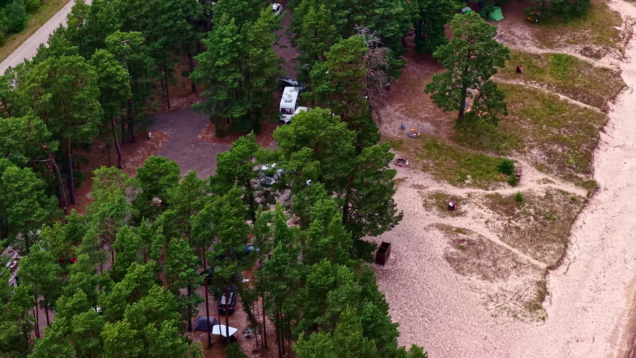 An aerial drone captures a beautiful summer day at a coastal campsite in Engure, Latvia, with motorhomes and camper vans parked in a pine forest right next to the sandy beach of the Baltic Sea