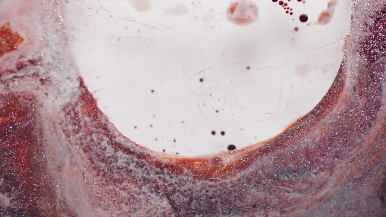A macro shot captures a white liquid added to red glitter ink. The inks swirl and separate, forming a textured, colorful surface