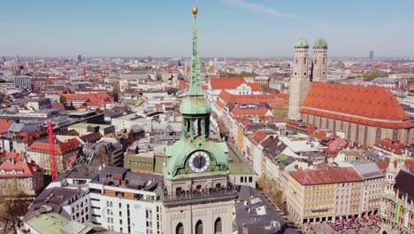 descending drone view of munich showing the green copper spire and clock face of st. peter’s church, frauenkirche’s twin domes, and the dense urban fabric of the bavarian capital under blue skies