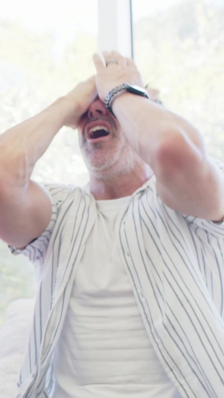 Exclaiming man in striped shirt covering face, upset, reclining on couch at home, partner watching