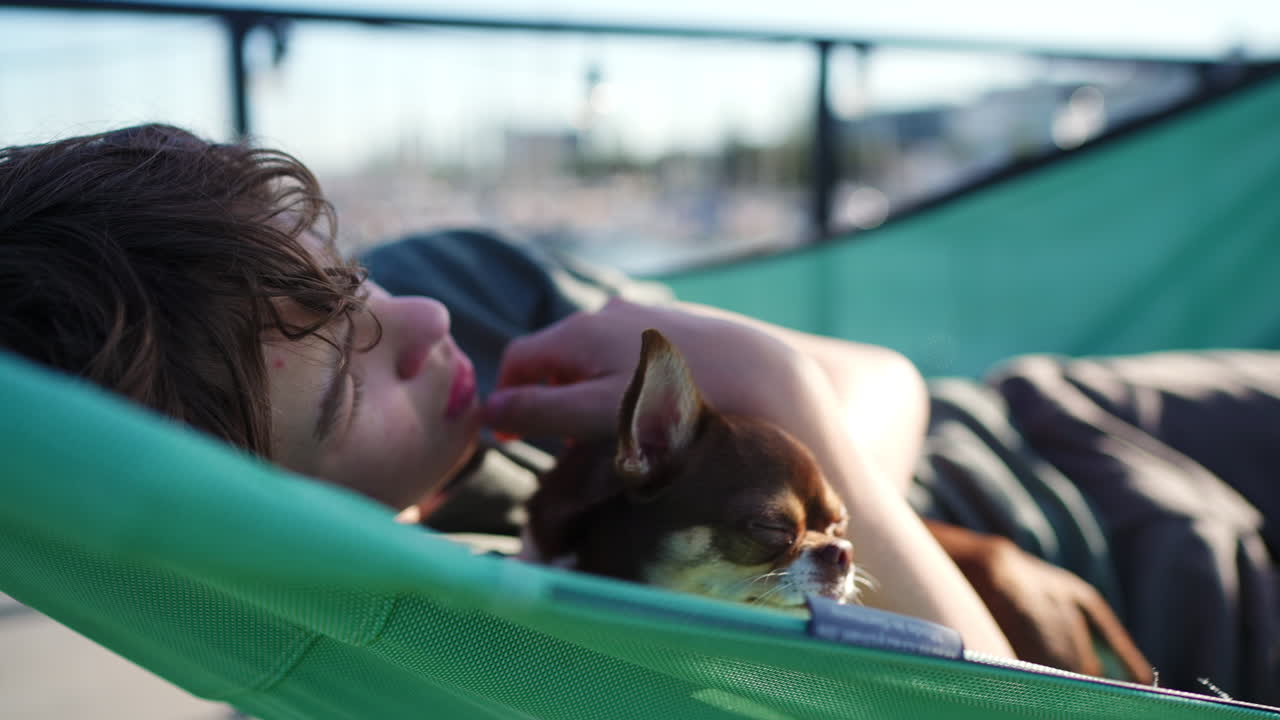 Young teenager lazily swinging in green hammock, sharing peaceful outdoor moment with small brown chihuahua, embodying carefree summer relaxation and deep companionship