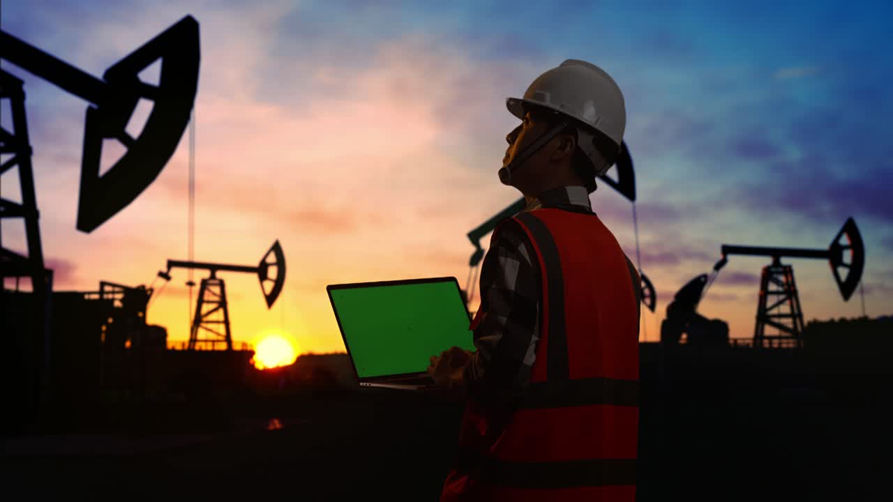 vista trasera de un ingeniero masculino asiático con casco de seguridad trabajando en una computadora portátil de pantalla verde y mirando a su alrededor mientras está de pie frente a las bombas de aceite, durante la puesta o el amanecer