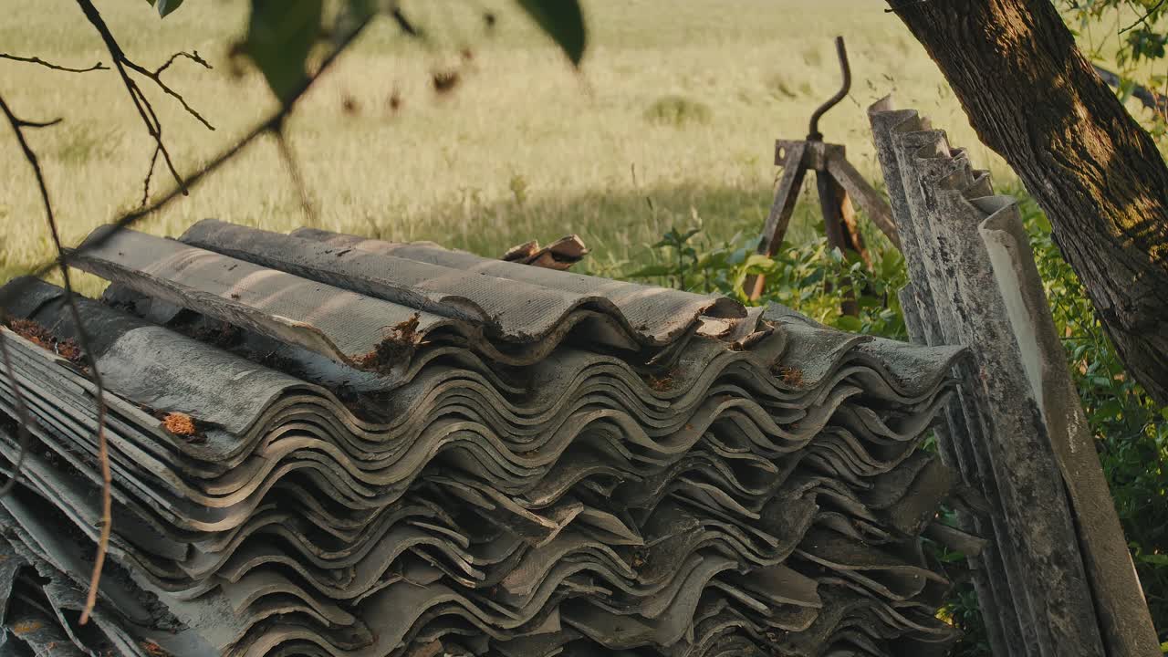 Man stacking old asbestos roofing tiles outdoors, performing manual labor in a rural setting. Close-up shot showing hands at work with hazardous construction material.
