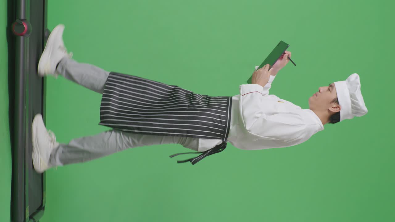 으로 보이는 아시아 남성 셰프가 녹색 스크린 배경 스튜디오에서 트레드밀을 는 동안 태블릿에 메모를 취하고 있습니다.