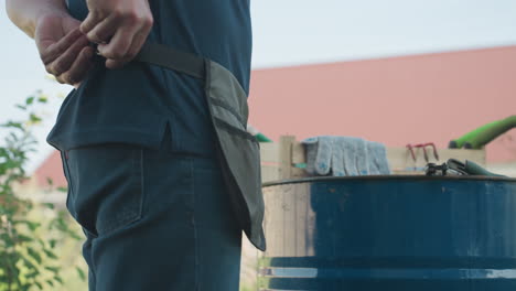 Close up man putting on apron belt around waist beside wooden crate on blue drum with gloves, trowel, fork, sprayer, yard house fence in background, organizing gear for garden work