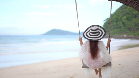 Mujer-Irreconocible-Con-Vestido-De-Verano-Y-Sombrero-Despojado-Columpiándose-En-Un-Columpio-De-árbol-Con-Cuerdas-En-Un-Asombroso-Fondo-Con-Vistas-A-La-Playa-En-La-Isla-De-Tailandia---Vista-Trasera-A-Cámara-Lenta