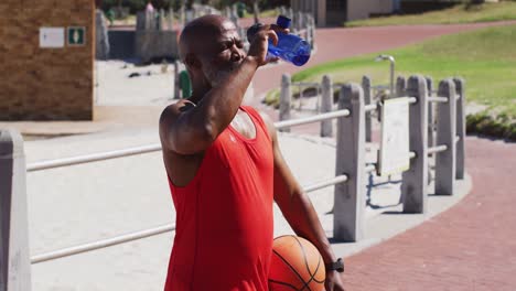 hombre afroamericano sénior con baloncesto bebiendo agua en la cancha cerca de la playa